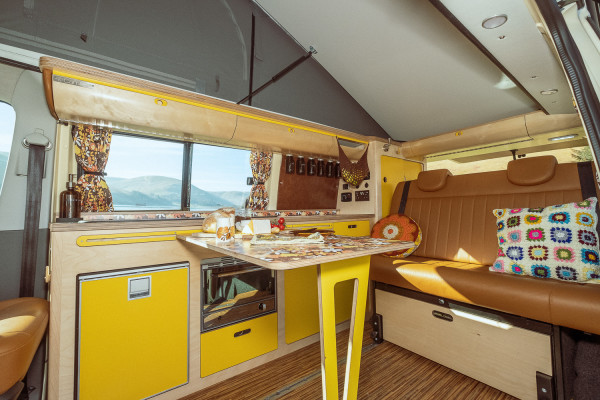 The interior of a retro-styled campervan showcases bright yellow cupboards, cheerful patterned curtains, and a brown leather sofa adorned with colourful crochet pillows. A pop-up table is set for a meal with mountains visible through the large window.