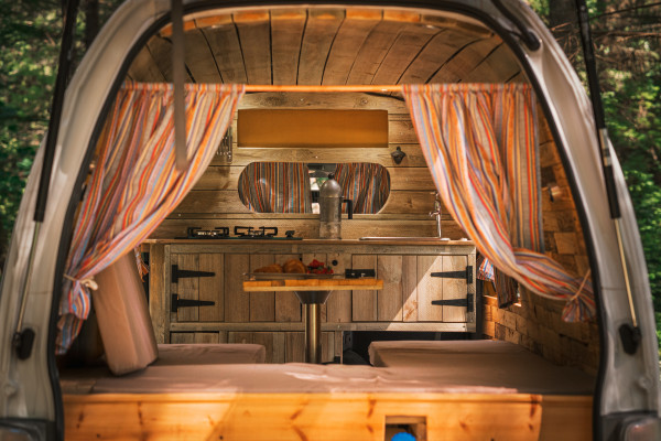 A cosy, rustic camper van interior with wooden panelling, striped curtains, and cushioned benches. A small kitchenette is visible with a hob, sink, and a wooden table holding fruit. The back doors are open, offering a view into the green, forested surroundings.