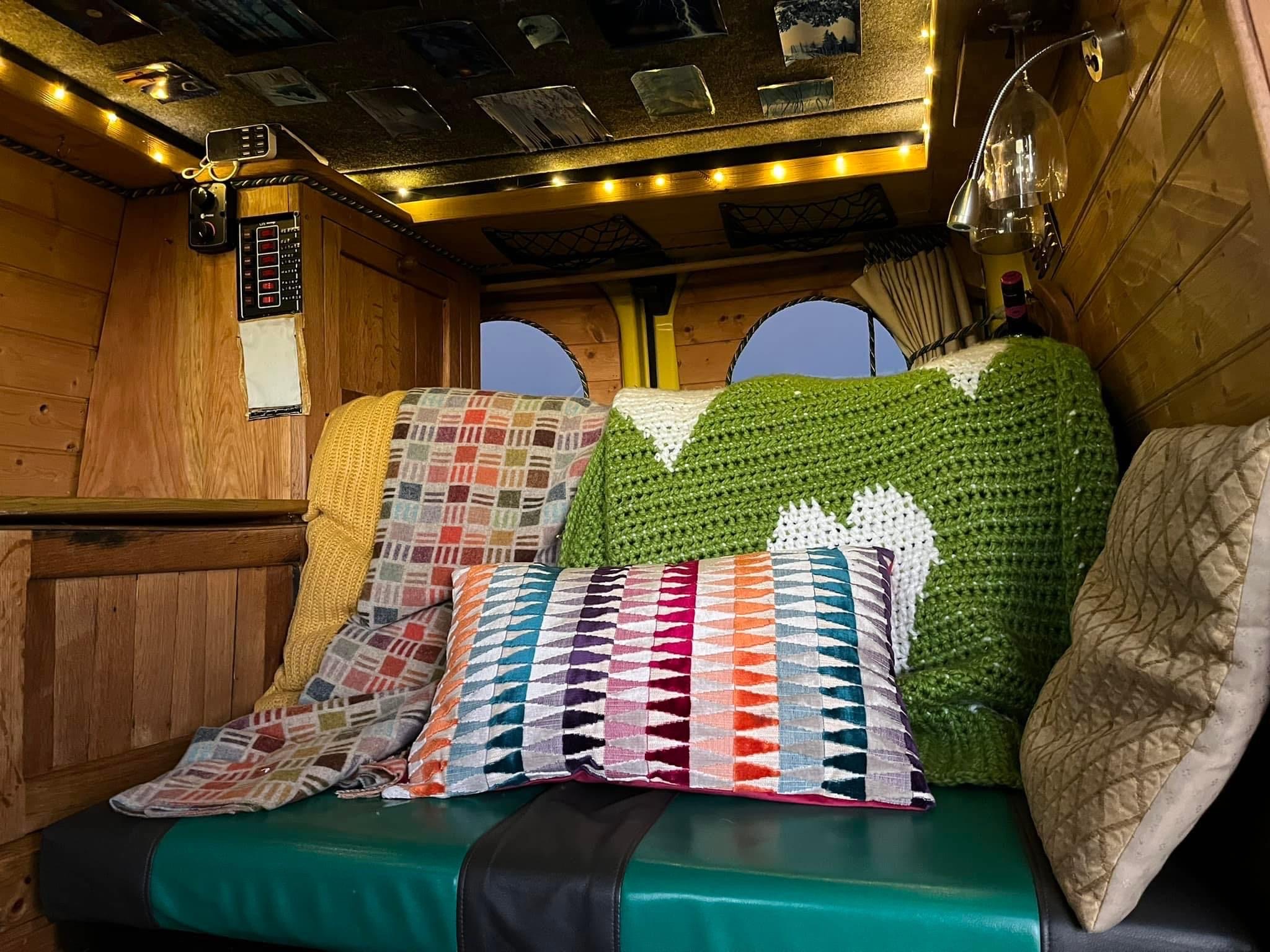 The image shows the cozy interior of a camper van. The seating area features a blue-green cushioned bench, draped with a green crochet blanket and a yellow quilt. A colorful geometric-patterned pillow rests on the bench. The wooden walls and soft lighting create a warm, inviting atmosphere.