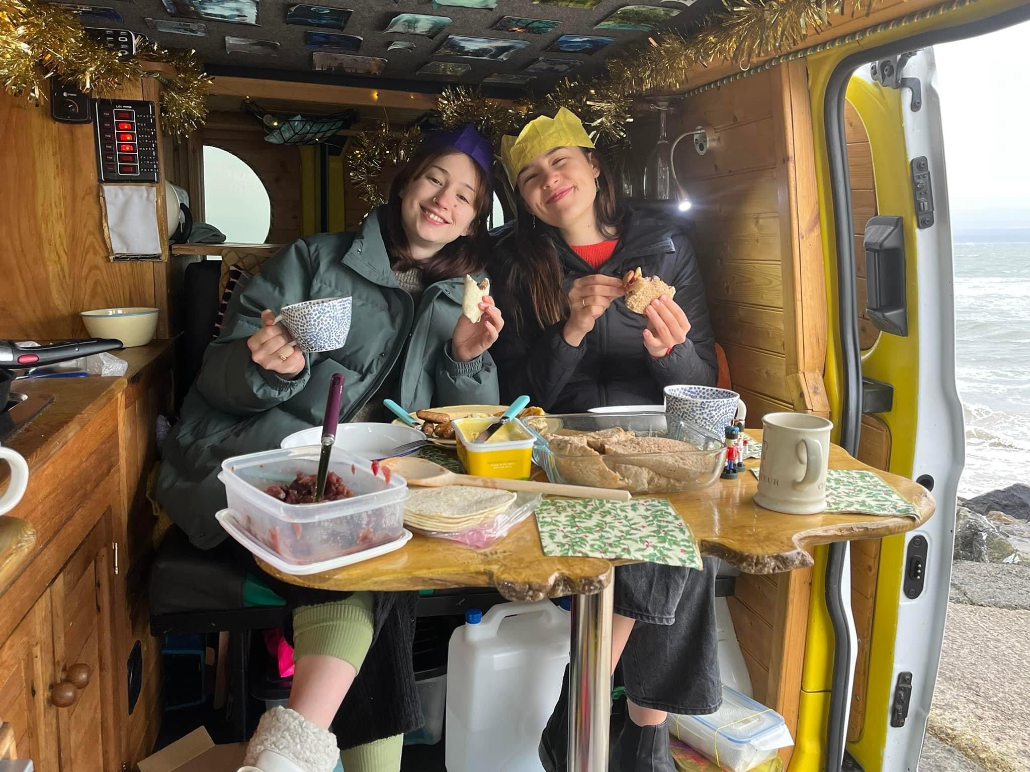 Two women sit inside a cozy, brightly decorated van by the seaside, smiling and enjoying a meal. They both wear colorful paper crowns and hold mugs and food. The van has wooden paneling, festive tinsel, and various containers of food on a small table. The ocean and rocks are visible outside.