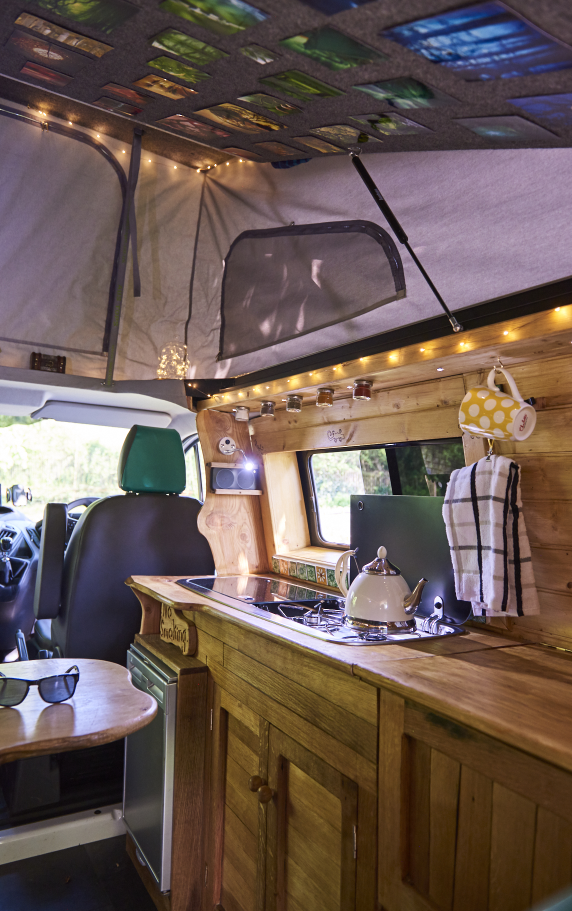 Interior of a camper van showcasing a compact kitchen. It features a stovetop with a kettle, a wooden countertop, spice jars mounted above, a checkered towel hanging, and a yellow polka-dotted mug. The van has green seat covers and is well-lit with both natural light from windows and string lights.