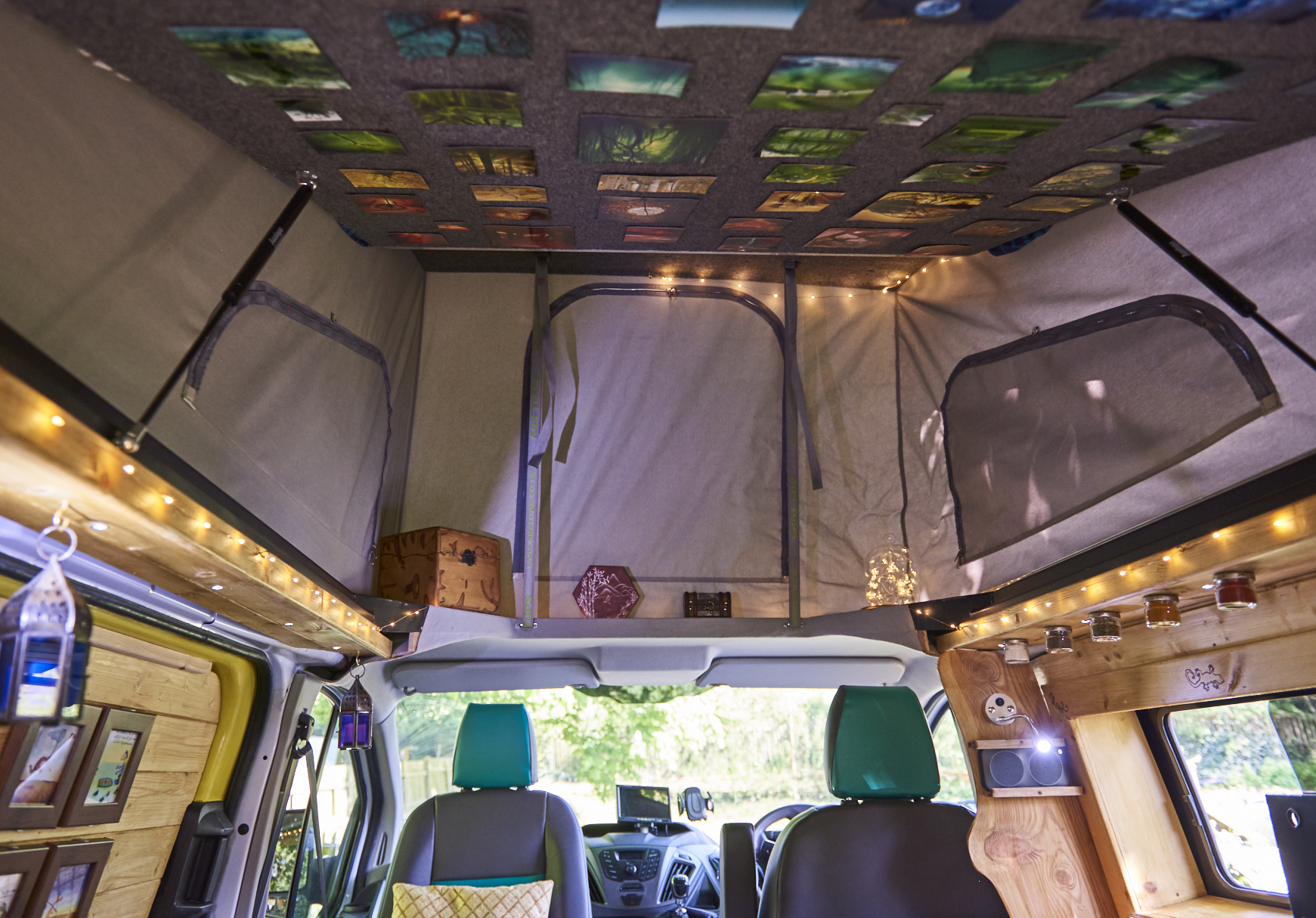 Interior of a camper van with cozy decorations. The ceiling is adorned with photographs. The walls are decorated with wooden accents and small jars. Fairy lights illuminate the space, providing a warm ambiance. The seating area includes green and black seats with pillows.