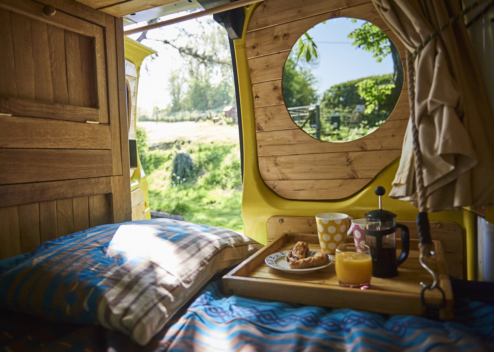 A cozy yellow campervan interior with wooden finishes. There's a bed draped with a blue blanket, and a tray on it that holds a French press, a glass of juice, and a polka-dotted mug. Through the large circular window, a sunny, green, grassy landscape with trees is visible.
