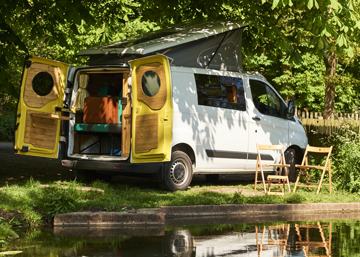 A white camper van with open yellow rear doors is parked by a lakeside, partially shaded by trees. Two wooden chairs are set up outside the van, facing the water. Inside, the van features a cushioned seating area and light wooden interior elements. It's a sunny, serene outdoor setting.