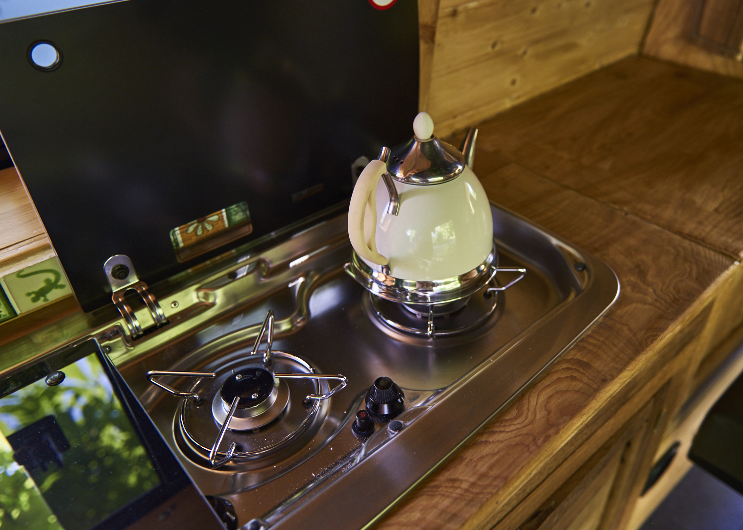 A compact kitchen setup inside a van features a stainless steel sink and a dual-burner gas stove. On one burner rests a vintage-style white and silver kettle. The surrounding wooden countertop has a natural finish, enhancing the cozy, mobile living space ambiance.