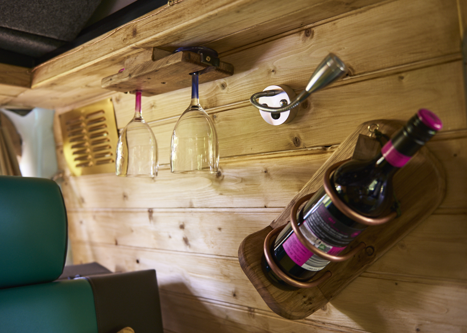 A close-up of a wooden interior wall of a camper van showcasing a compact wine storage solution. Two wine glasses hang upside down from a wooden rack, and a wine bottle is secured in a wall-mounted wooden holder. The background features light-colored wooden paneling and a metal hook.