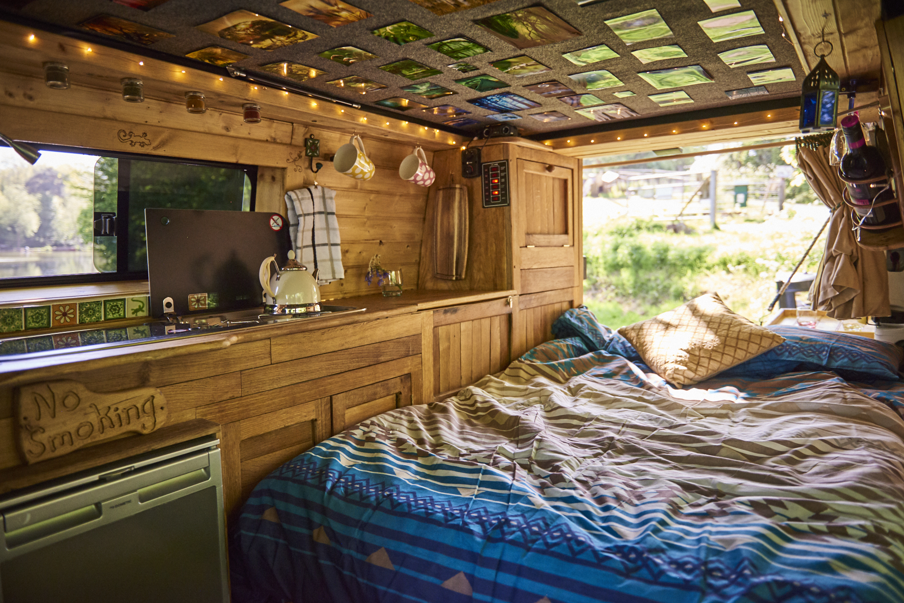 Interior of a cozy, wooden camper van. The bed, covered with a blue and white patterned blanket, is adjacent to a kitchenette with a small stove, teapot, and various utensils. The ceiling is decorated with colorful tiles. The window above the bed reveals a scenic, sunlit view of trees and grass.