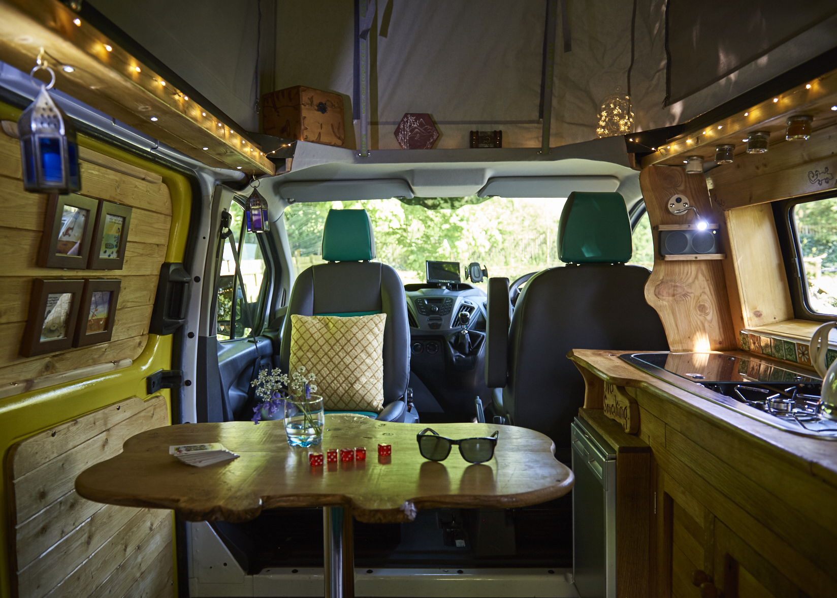 Interior of a cozy, well-lit camper van. The wooden table holds sunglasses, a candle, red dice, and a small vase with flowers. The front seats are upholstered in gray and teal. The cabinets and walls are wooden, adorned with string lights, small pictures, and miscellaneous items. Trees are visible through the front windshield.
