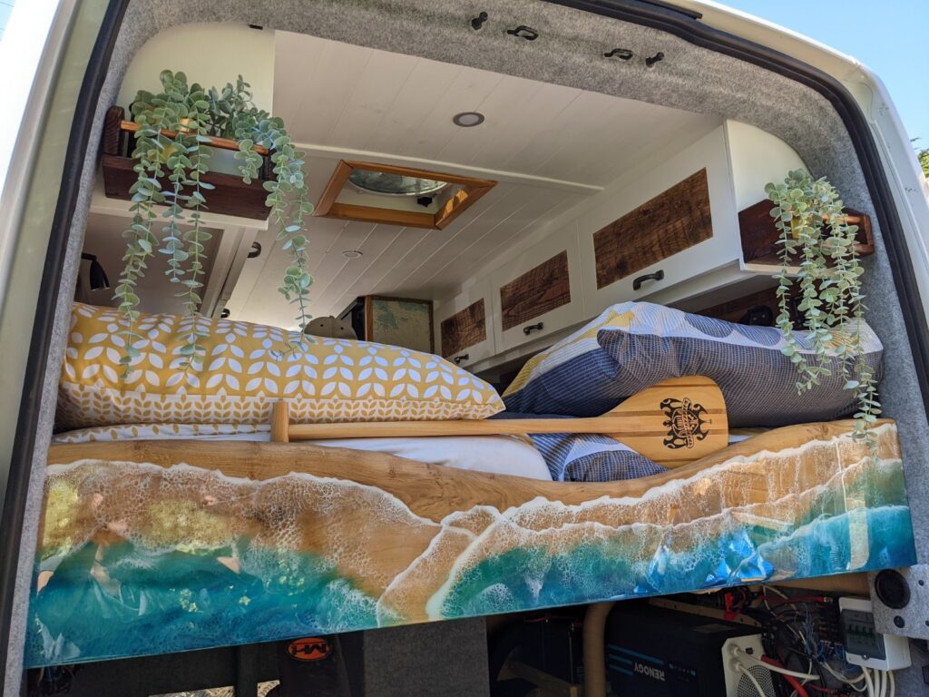 The interior of a camper van is shown from the rear with a bed taking up most of the space. The bed has colorful bedding, including a pillow with a geometric pattern and a comforter featuring an ocean wave print. Hanging plants decorate the upper shelves, adding a touch of greenery.