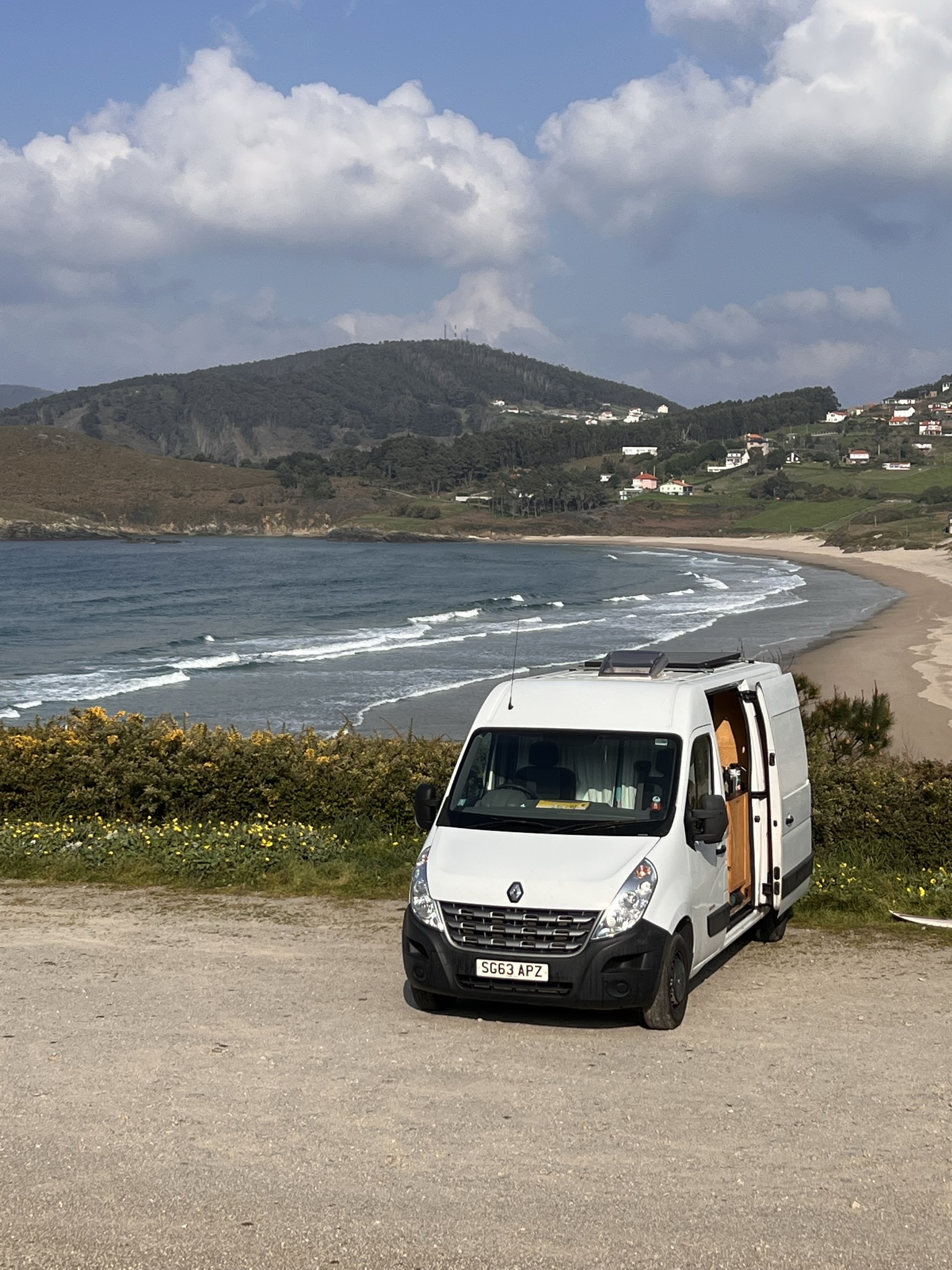 A white camper van with an open sliding door is parked on a grassy area overlooking a curving sandy beach and the ocean. Waves crash gently on the shore. In the background, green hills and scattered houses dot the landscape under a partly cloudy sky.