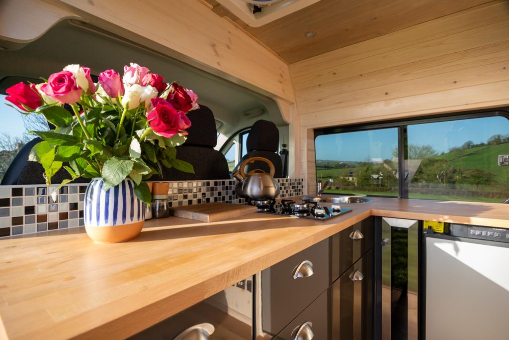 A modern camper van kitchen features a light wooden counter, a stainless steel kettle on a stovetop, a cutting board, and a vase of colorful roses. The interior has wood-paneled walls and a window revealing a scenic view of green fields. Dark cushioned driver and passenger seats are visible.