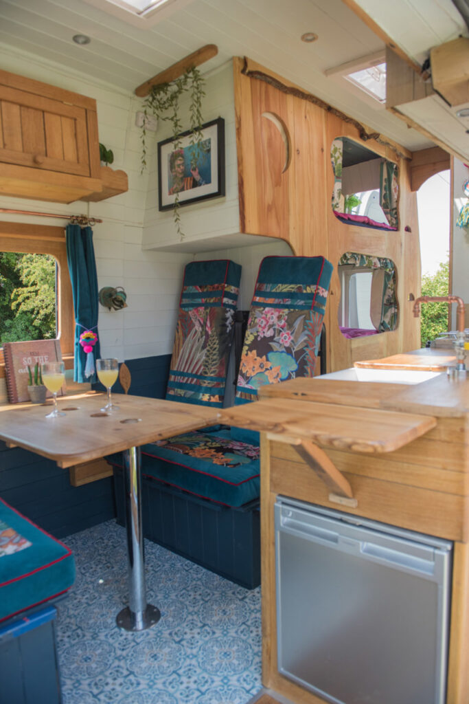 Interior of a cozy camper van with colorful patterned cushions, a wooden fold-out table, and two glasses of wine on the table. A small vase with flowers adds charm to the setting. The right side features a wooden structure with windows that let in natural light, enhancing the warm atmosphere.