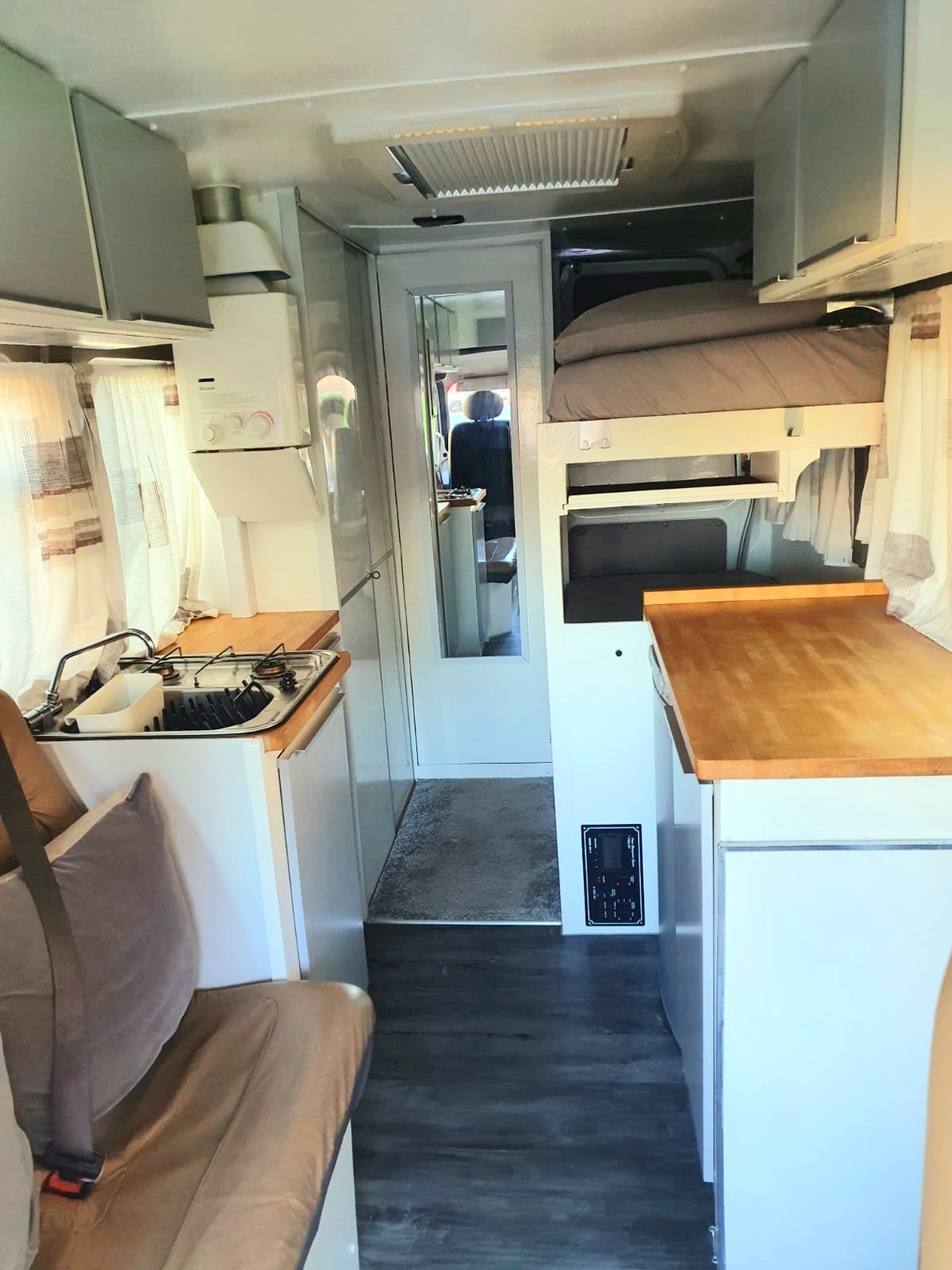 Interior of a campervan showing a small kitchen area on the left with a stove and sink, a bed on a raised platform on the right, a wooden countertop below the bed, and a walkway leading to the back of the van. Light flows through the windows, illuminating the cozy space.