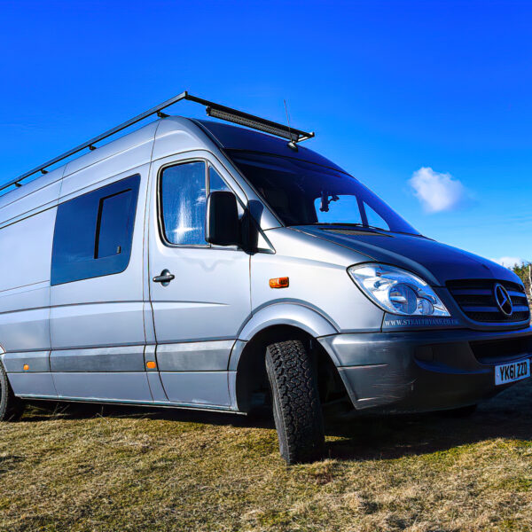 A silver van with a roof rack is parked on a grassy area under a clear blue sky. The van is partially off-road with one front wheel on higher ground. In the background, there are scenic mountains and patches of trees. The image suggests an adventurous, off-grid setting.