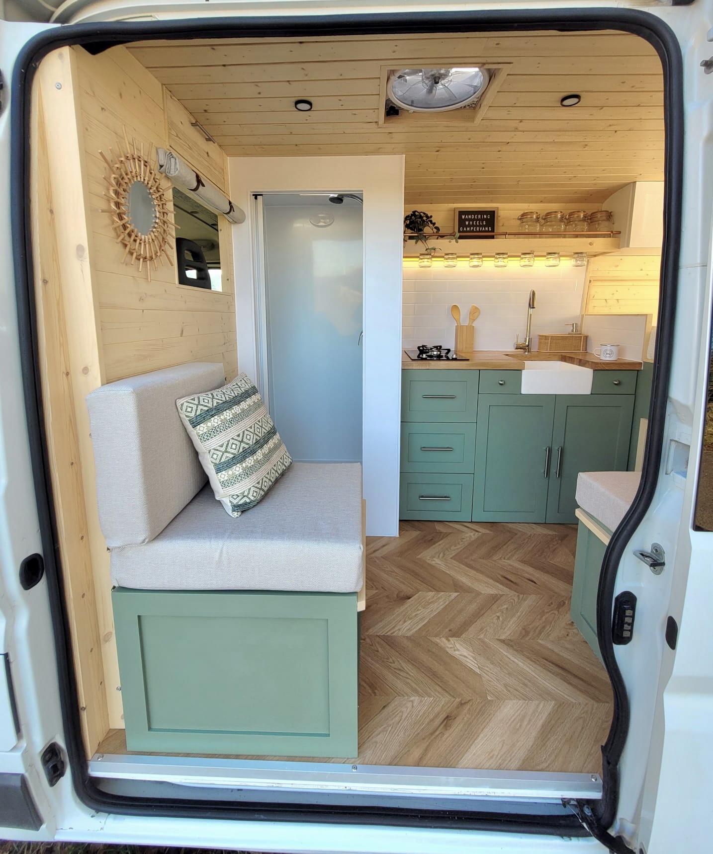 Interior of a converted camper van showcases a cozy living space featuring a built-in cushioned bench with decorative pillows on the left. The right side holds a green kitchenette with drawers, a farmhouse sink, and floating shelves, all under warm LED lighting. The floor is herringbone-patterned wood.