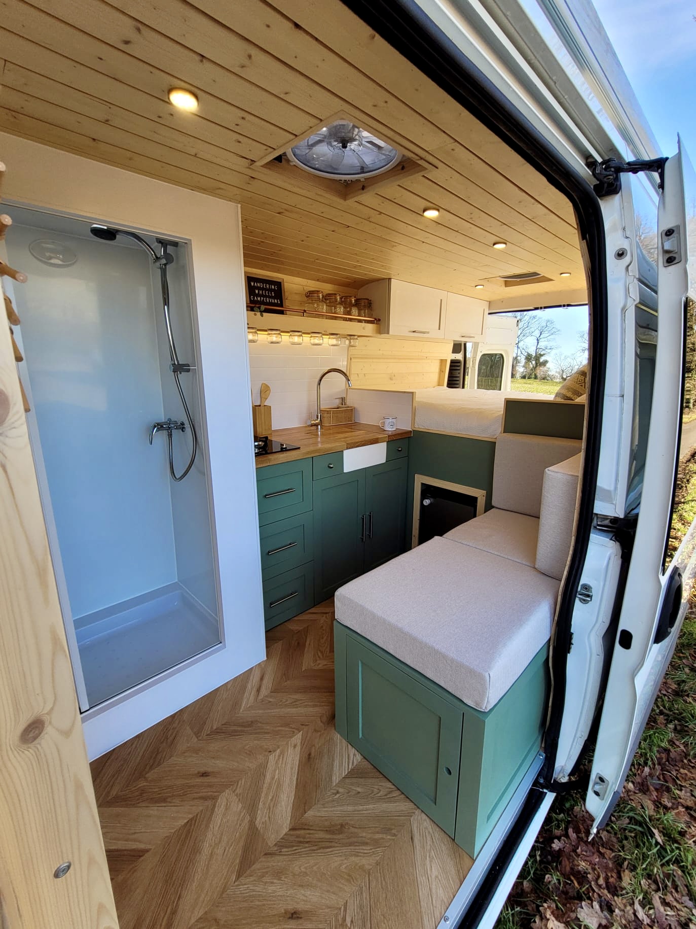 Interior of a converted van showcasing a cozy living space. On the left, there's a white shower with a glass door. The middle has a kitchen area with green cabinets, a sink, and wooden countertop. On the right, a seating area with beige cushions. A bed is visible above the seating area.