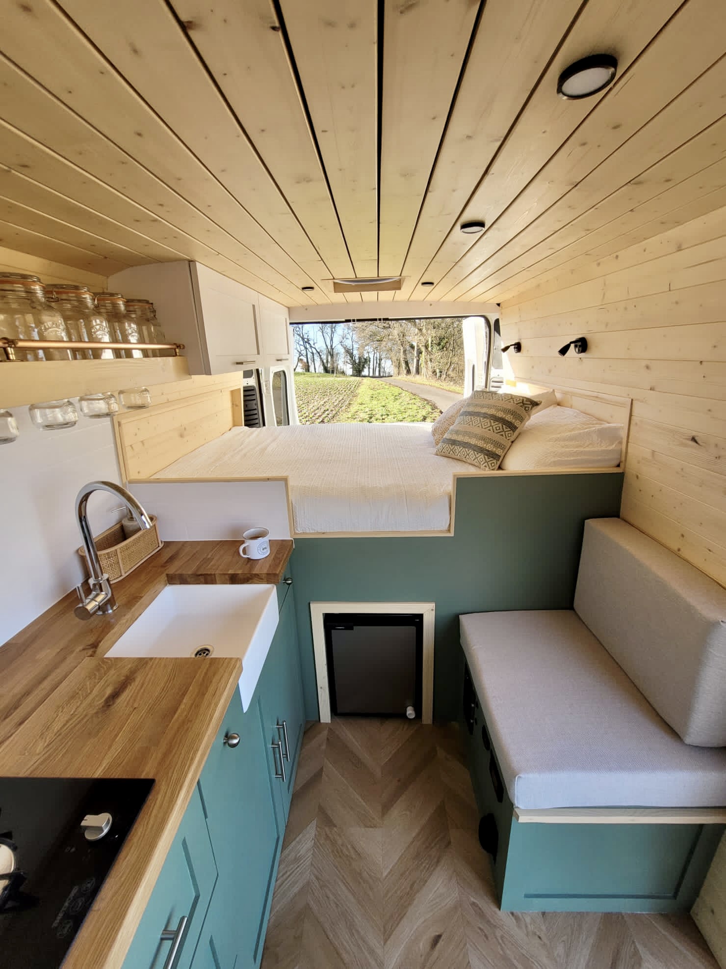 Interior of a minimalist camper van featuring a cozy bed elevated above a bench seat, adjacent to a small kitchenette. The kitchen area includes a white sink, wooden countertops, and glass jars on a shelf. The walls and ceiling are lined with light wooden paneling, creating a warm, inviting atmosphere.