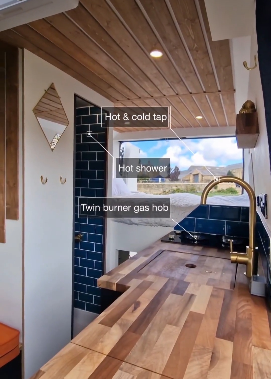 Image of a tiny house kitchen and bathroom area. Features include a hot and cold tap over a wooden countertop with a twin burner gas hob. There's a hot shower with blue tiles in the background and a mirror. A large window reveals a scenic outdoor view of a partly cloudy sky and green landscape.
