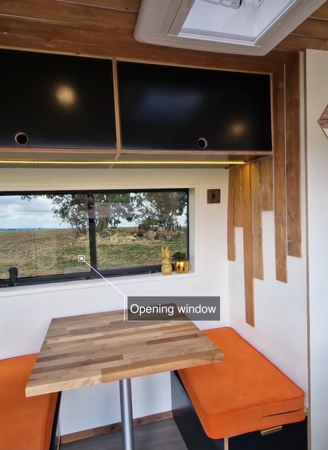 Interior of a cozy RV featuring a wooden table with two benches, each covered with orange cushions. Above the table is a large window labeled