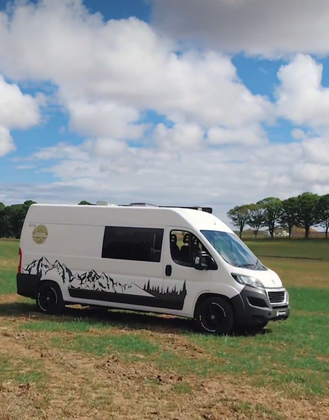 A white camper van is parked on a grassy field under a cloudy blue sky. The van has a black silhouette graphic of mountains and trees along its lower side. Behind the van, there are scattered trees on the horizon. The scene conveys a peaceful, outdoor adventure vibe.