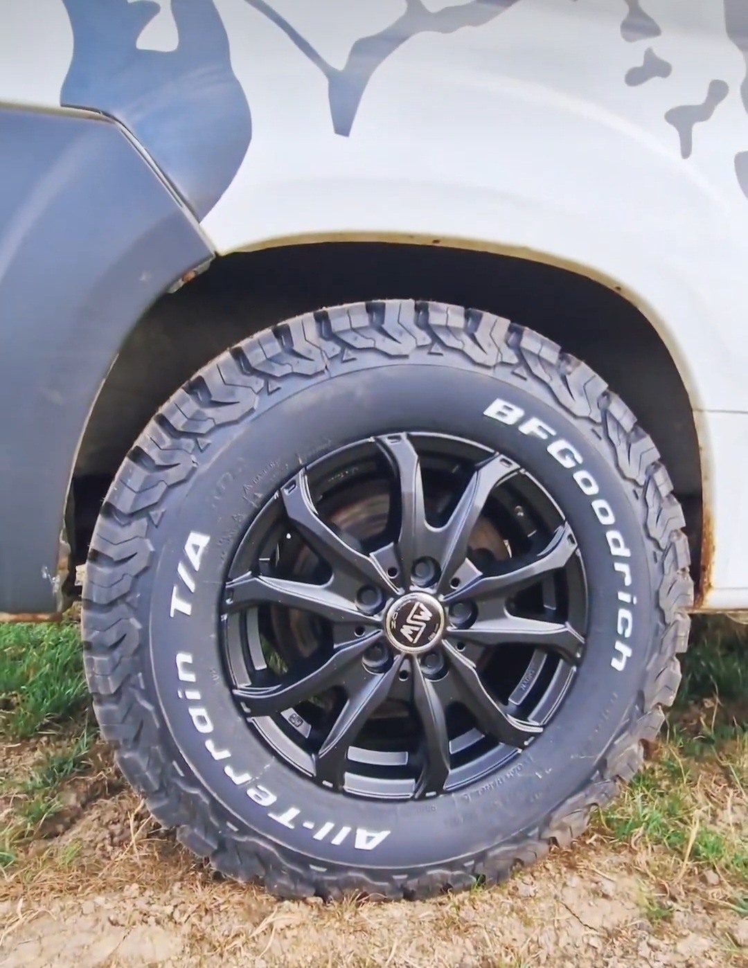 Close-up image of the rear wheel of a white off-road vehicle with a camouflage pattern. The wheel features a chunky BFGoodrich All-Terrain T/A tire and a black multi-spoke rim with a metallic logo at the center. The vehicle is parked on a grassy terrain with dirt visible on the tire tread.