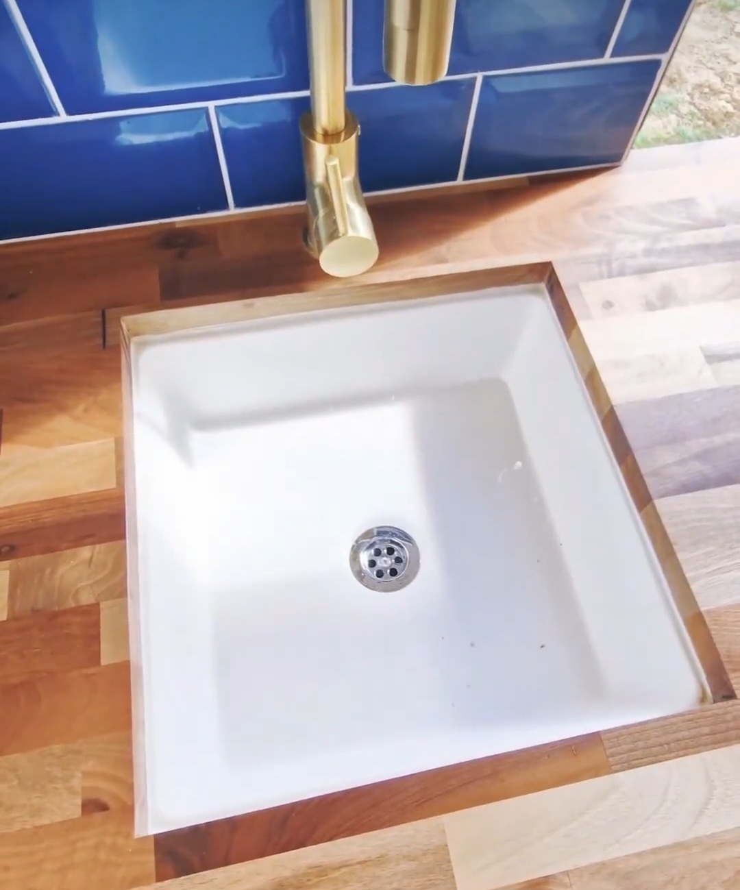 A square white sink is set into a wooden countertop. Above the sink is a brass-colored faucet, partially visible. The countertop features various shades of brown wood planks, adding a rustic feel to the modern design. Blue tiles are just barely visible at the top edge of the image.