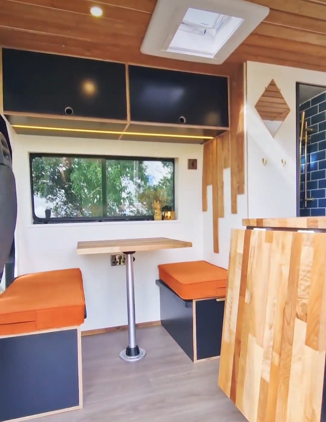 A cozy and modern tiny home interior featuring a wooden table with two orange cushioned benches on either side, set against a large window. Black cabinets with wood trim are above the window, and a skylight brightens the space. Wood accents and greenery are visible through the window.