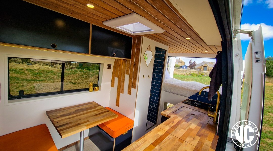 Interior of a modern camper van with wood accents. The ceiling and countertops are wooden, and there are black storage cabinets. A dining area has orange cushions and a large window. A cozy bed with white linens is in the background, and a small skylight provides additional light.