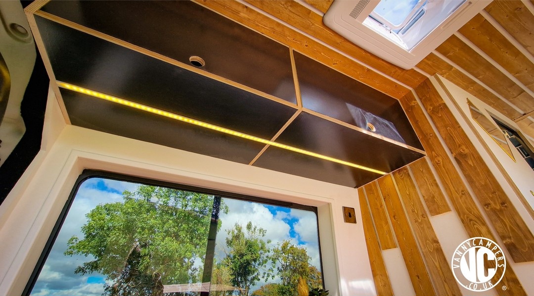 The image shows the interior of a modern tiny home. A large window offers a view of a tree and blue sky with some clouds. Above the window, there are black, open shelving units with built-in yellow lighting. The walls feature a mix of white paneling and wood accents. The logo