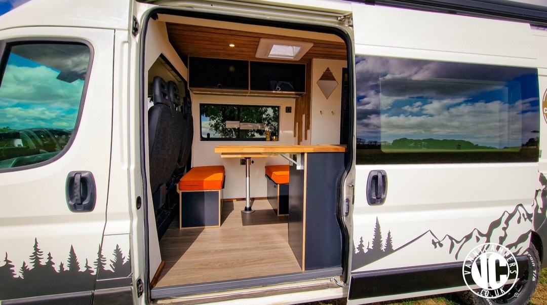 The image shows the interior of a modern camper van with the sliding door open. Inside, there's a wooden table with two cushioned benches on either side, featuring bright orange cushions. The floor is wooden, and large windows allow natural light, revealing an outdoor landscape with trees and mountains painted on the van's exterior.