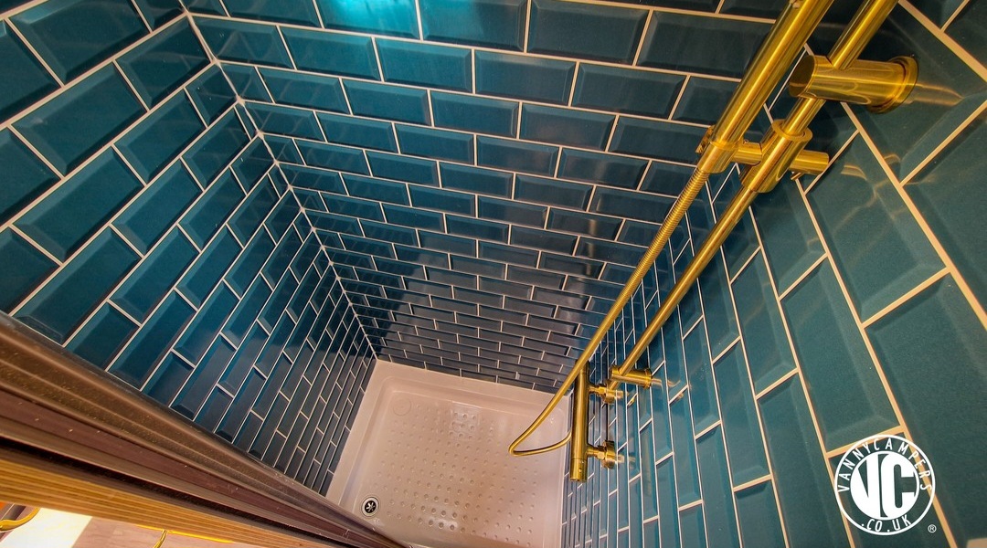 The image shows a shower stall with vibrant teal subway tiles covering the walls in a brick pattern. The showerhead and fixtures are brass, adding a touch of elegance. The floor of the shower is white with a slightly raised texture for grip. The corner reveals a small, enclosed, intimate space.