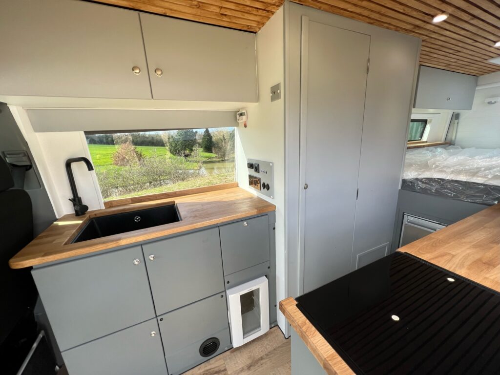 The image shows the interior of a modern camper van kitchen. It features gray cabinets, a black sink, a wooden countertop, and a window above the sink providing a scenic view of greenery. The ceiling has wooden paneling. On the right, there is a countertop with an induction cooktop.