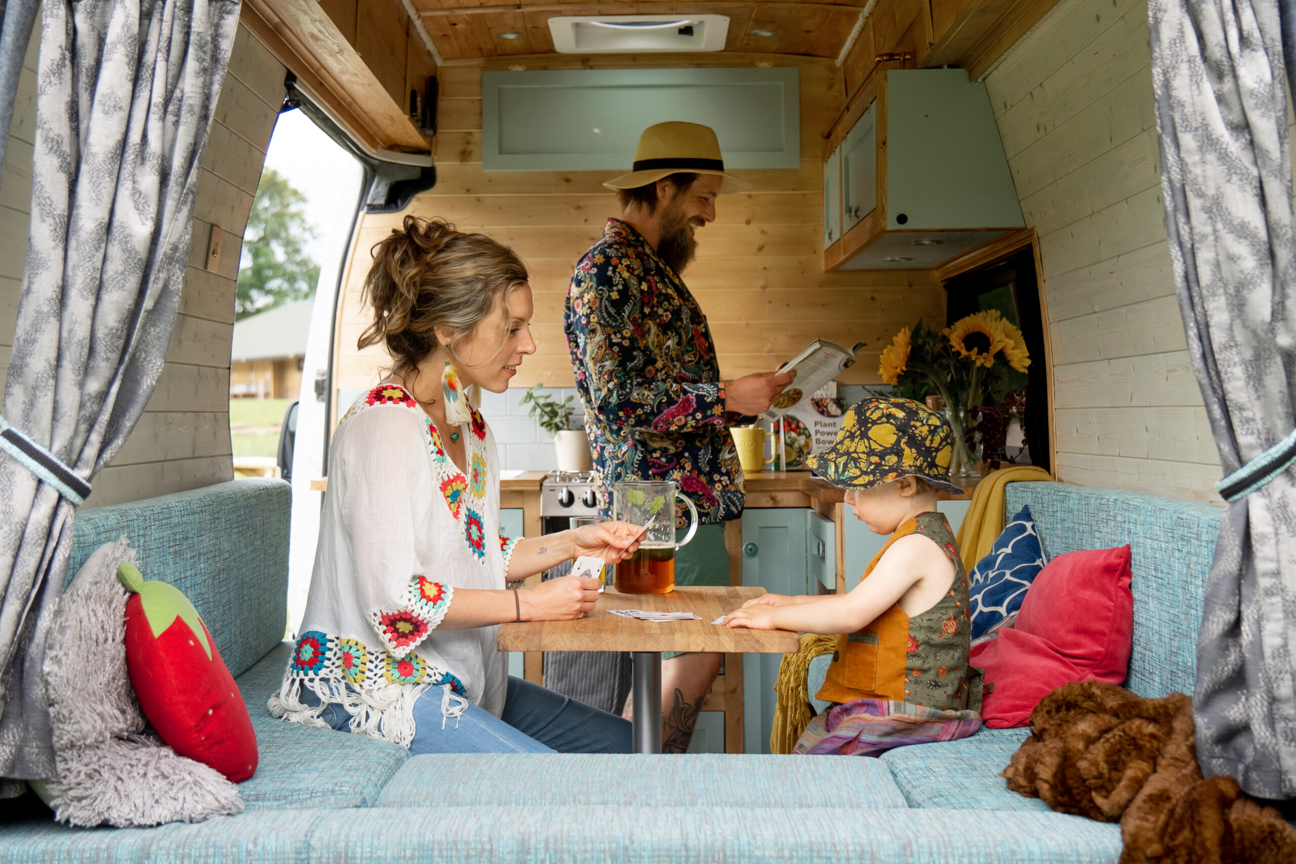 A family is gathered inside a campervan. A woman with a braided bun and colorful attire plays cards with a child who sports a patterned bandana and vest. A man with a beard and hat reads a book near the kitchenette. The interior features turquoise seating, wooden decor, and sunflowers.