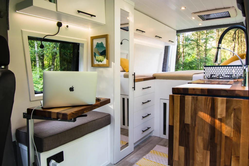 The interior of a stylish camper van features white cabinetry, a wood counter, a cushioned bench, and a laptop on a small wooden fold-out desk. A mirror-fronted storage unit with drawers sits against the wall, and a bed is visible at the back. Large windows offer a view of greenery outside.