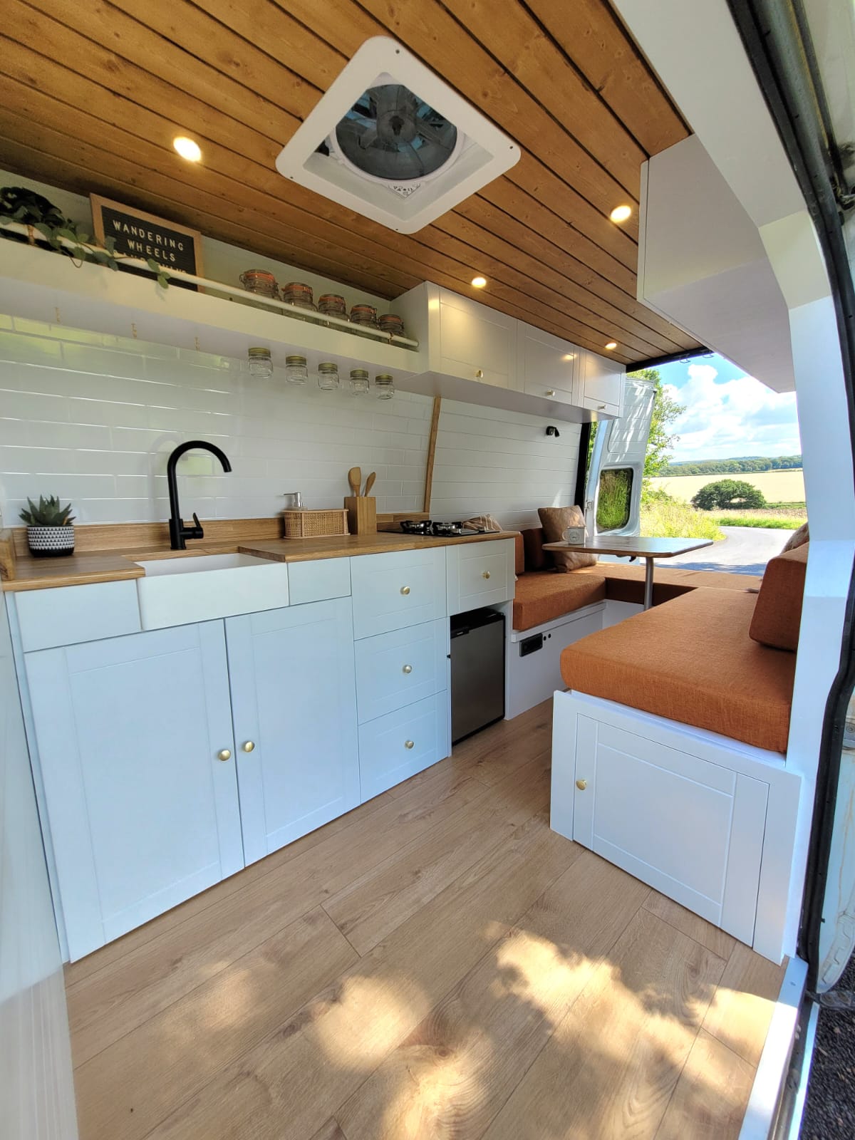 A modern, minimalist camper van interior features white cabinetry, a wooden countertop, and a sink with a black faucet. Open shelves hold jars near the ceiling. Orange cushioned seating lines the right side. A large window on the right offers a view of lush green fields and blue skies.