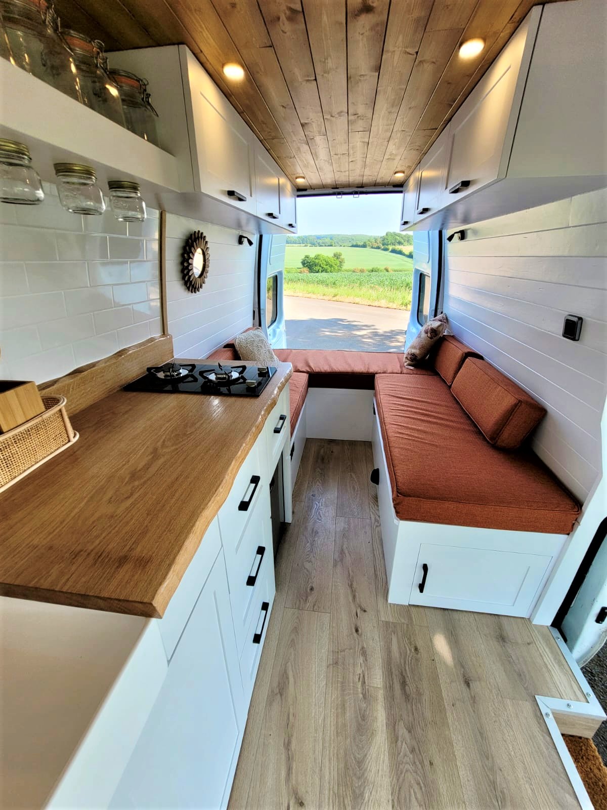 The interior of a neatly designed van conversion, showcasing a cozy, wood-accented kitchenette with a gas stove on the left and a narrow aisle leading to a seating area with orange cushions on the right. Large windows display a scenic view of a lush, green landscape outside.