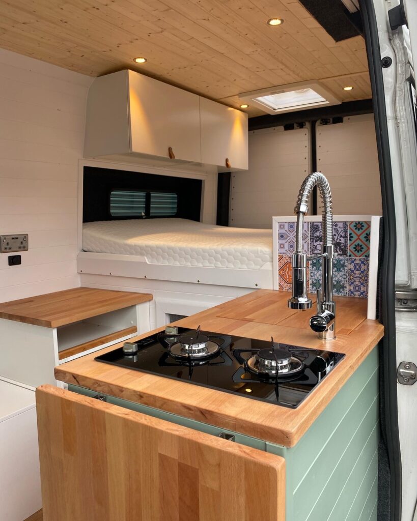 Interior of a modern camper van with wooden countertops. The foreground shows a compact kitchen area with a gas stove, sleek faucet, and colorful tiled backsplash. In the background, there is a neatly made bed with white bedding, and overhead cabinets for storage.