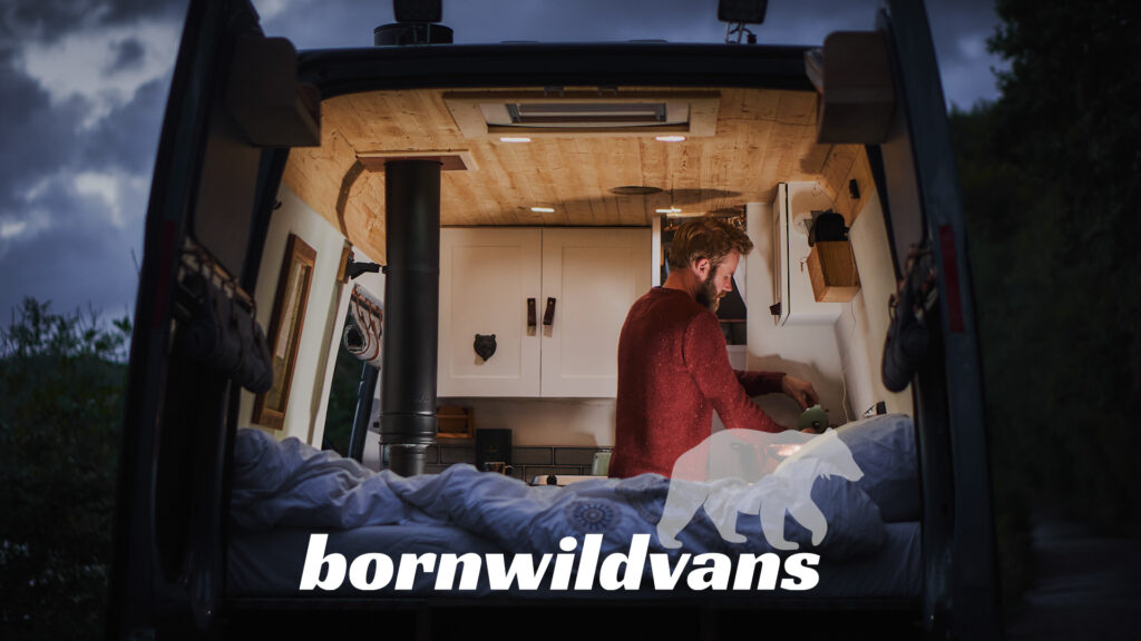 A person stands inside a cozy, well-lit van, which appears to be converted into a living space. The walls are white with wooden cabinets, and the ceiling is also wooden. The van doors are open, revealing a plush bed. The text "bornwildvans" and a bear graphic are prominently displayed at the bottom.