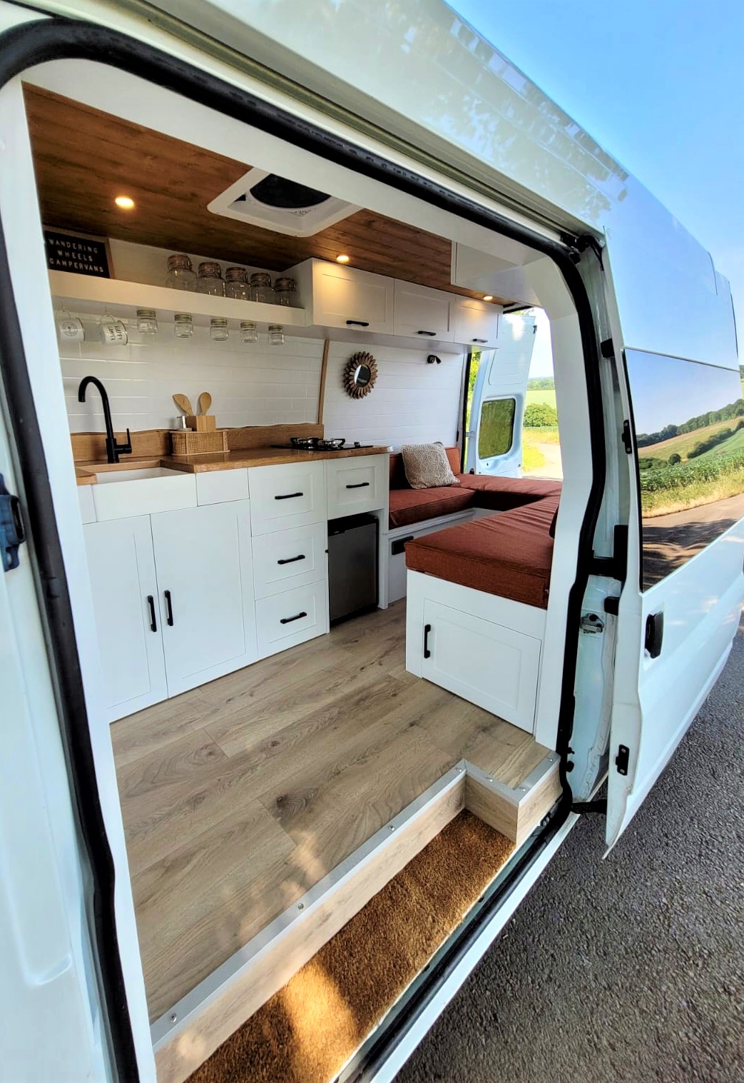 The image shows the interior of a white camper van converted into a cozy living space. It features white cabinets and drawers, a small sink with a black faucet, a wooden countertop, a small stove, a mini-fridge, and a cushioned seating area with a table. The back doors are open, revealing a scenic view outside.