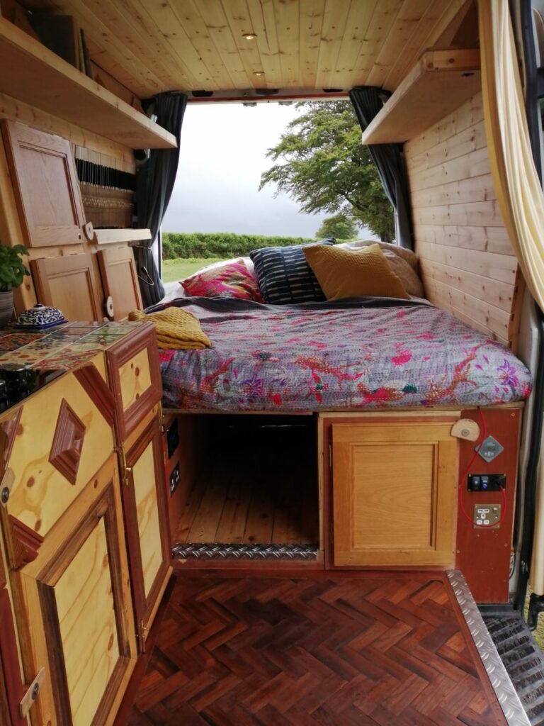 A cozy interior of a camper van. The van is equipped with a comfortable bed adorned with colorful pillows and a floral bedspread. Wooden cabinets and drawers in various patterns are built into the walls. Curtains hang beside the bed, and a scenic view of greenery is visible through the open rear doors.