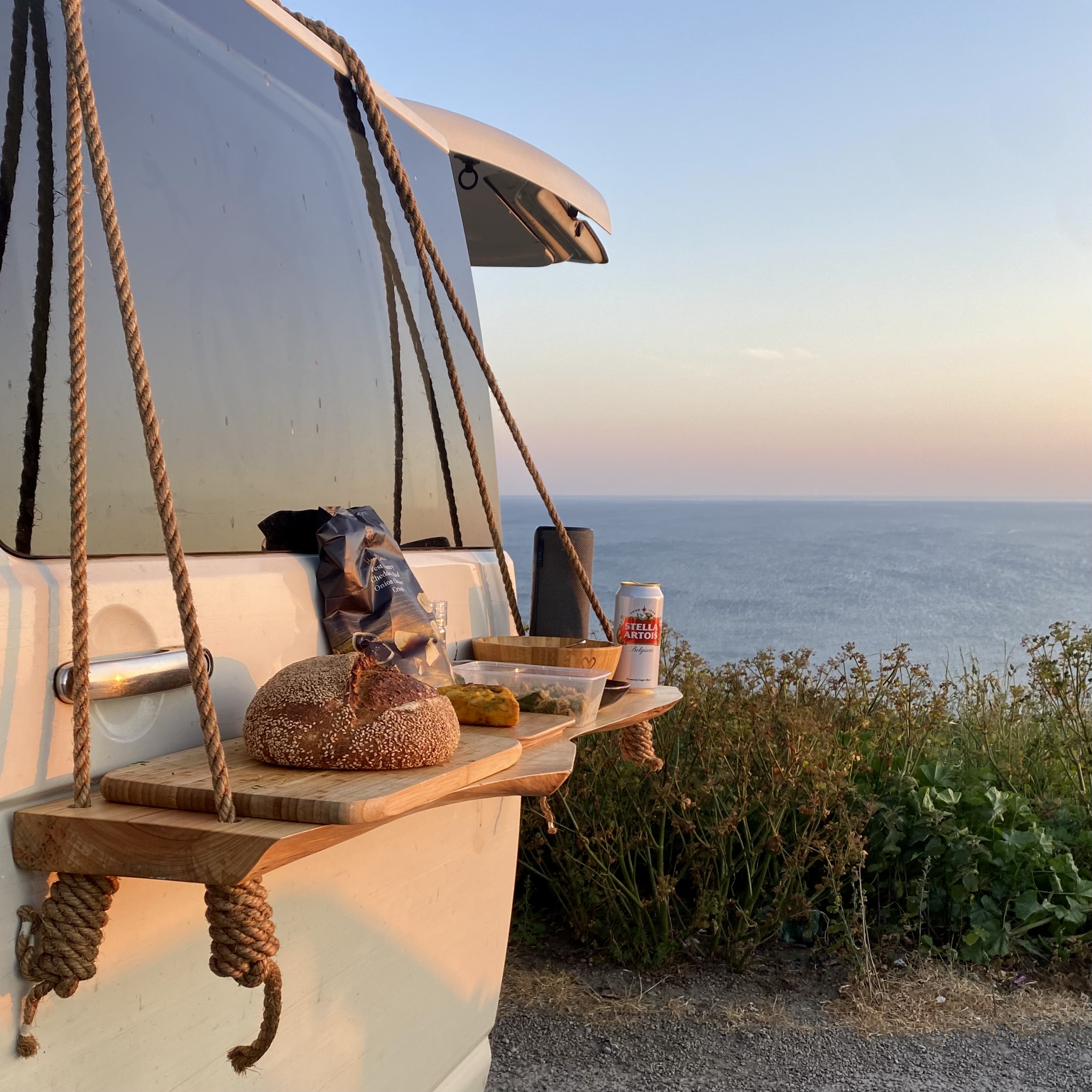 A mobile wooden table hangs from the rear door of a white van by thick ropes, set against a scenic ocean view at sunset. On the table are a whole loaf of bread, a jar of pickles, a bag of chips, a beverage can, and a plastic container. The surrounding vegetation adds a natural touch.