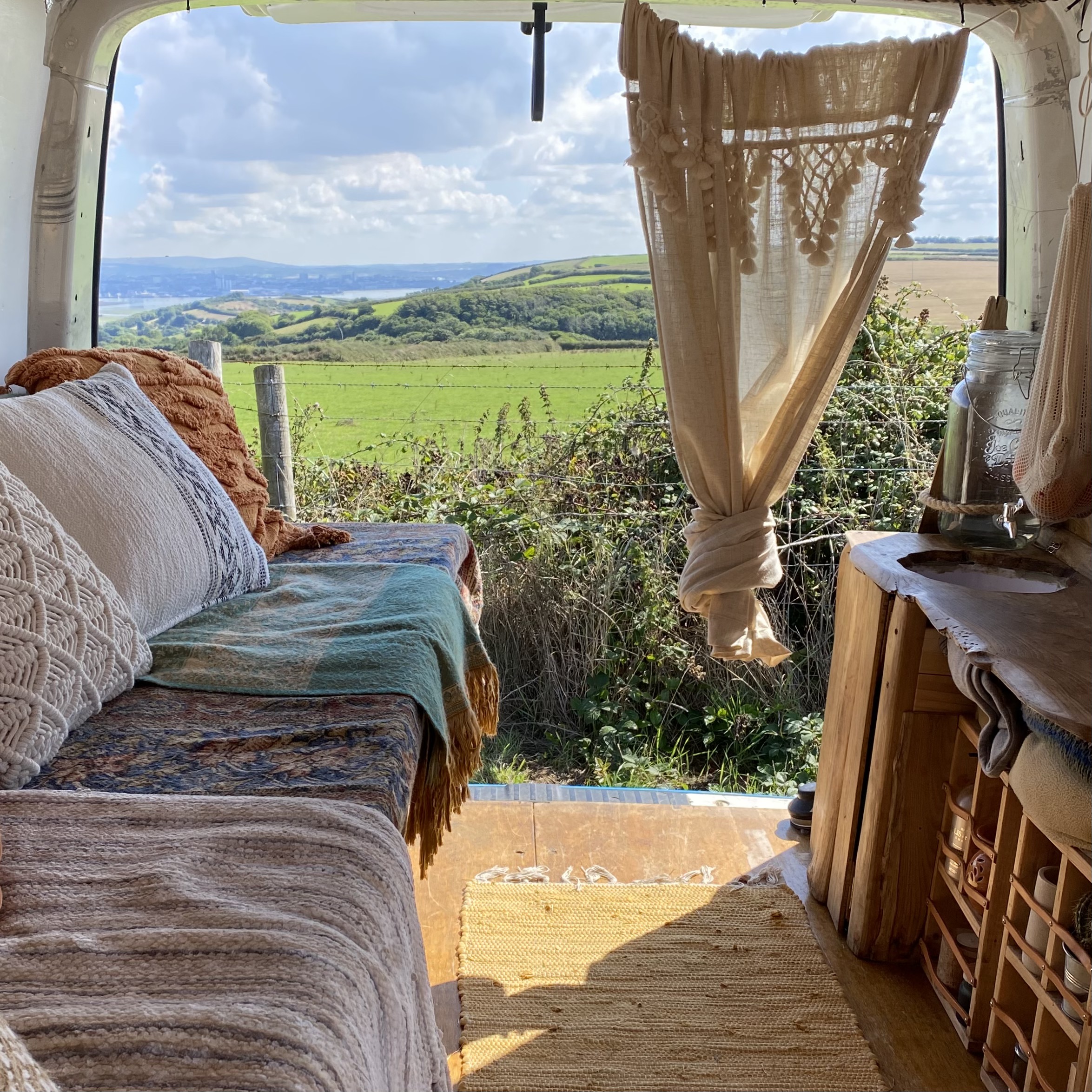 A cozy interior of a van converted into a tiny home with a colorful, patterned blanket on a bench seat, white and brown pillows, and sheer, knotted curtains. Through the open doors, a scenic view of lush green fields, rolling hills, and a bright blue sky can be seen.