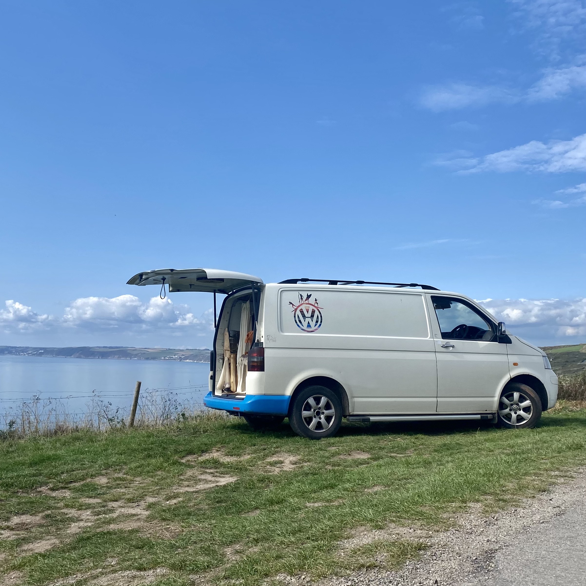 A white van with a blue lower trim and a VW logo is parked on a grassy hill overlooking a large body of water and distant land. The van's rear door is open, revealing curtains inside. The scene is under a bright blue sky with a few clouds.