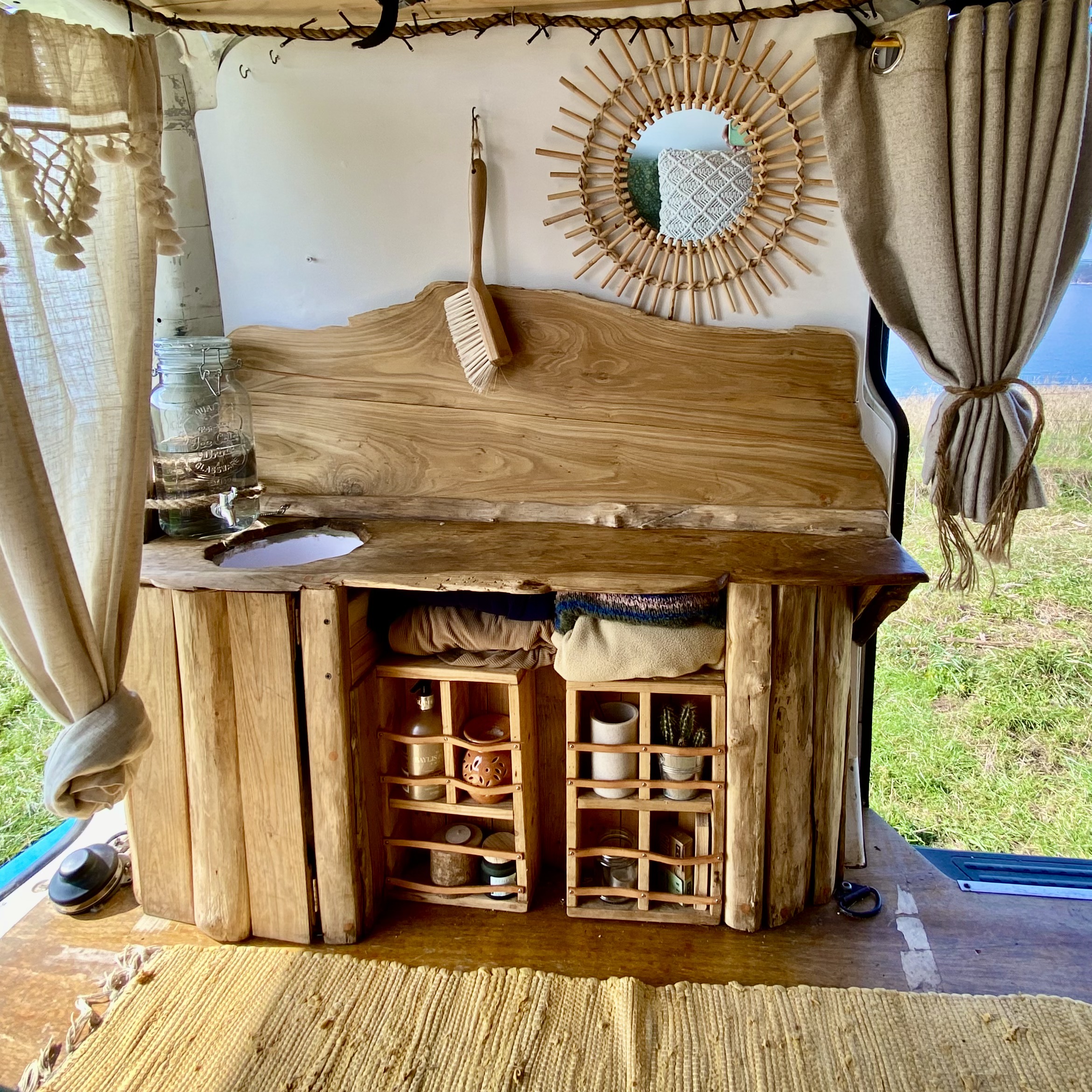 The image shows a rustic wooden countertop in a van with a built-in storage area. Various personal items are neatly organized on shelves and inside crates. A small round mirror with a sunburst frame hangs on the wall above the counter. Curtains are drawn open, and a woven rug covers the floor.