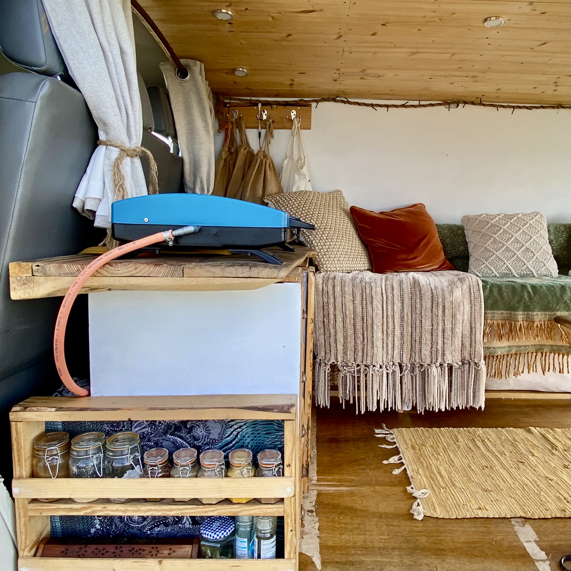 A cozy van interior features a compact, rustic kitchen setup. A blue portable stove sits atop a wooden counter with a gas hose attached. Below, jars filled with various items are neatly arranged. In the background, a cushioned seating area with textured pillows and a throw blanket is visible.