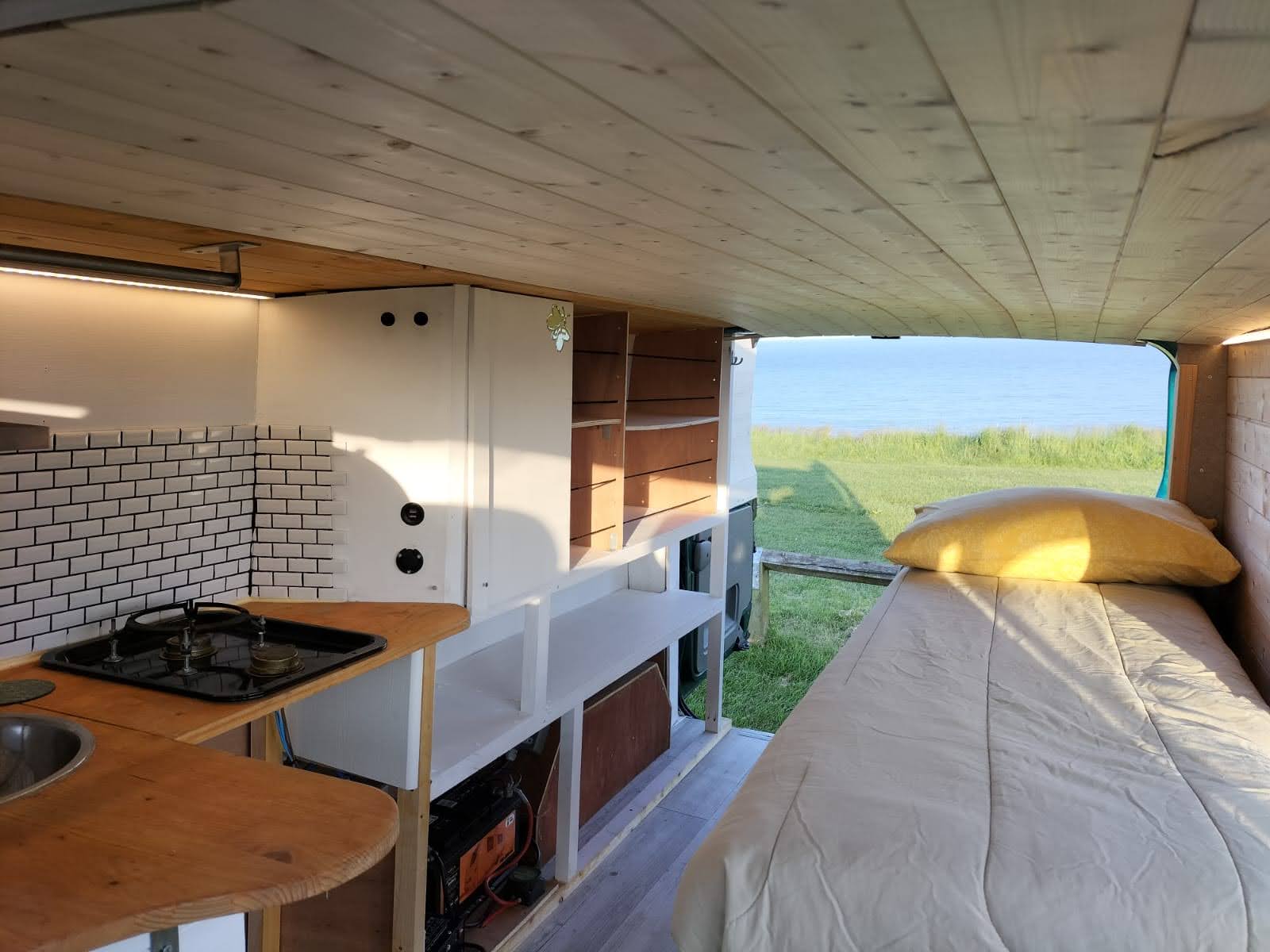 Interior of a cozy camper van converted into a tiny home. On the left, a small kitchen area features a wooden countertop with a gas stove and white tiled backsplash. Below are shelves and storage compartments. To the right is a bed with pillows, next to a large window revealing a grassy landscape and lake.