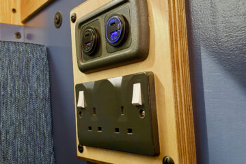 A wooden panel on a wall houses a set of electrical outlets. The top section features two circular USB ports with an illuminated blue display. Below, there is a standard UK double socket with two switches. The wall around the panel is partially covered in blue fabric and painted wood.