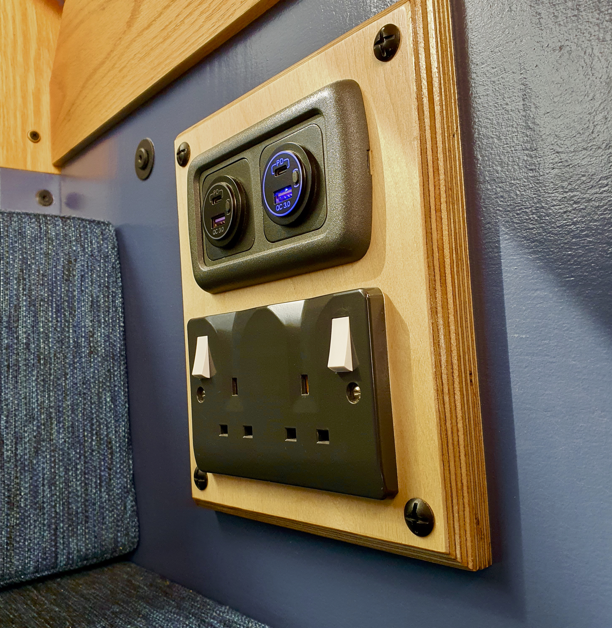 A wooden panel on a wall houses a set of electrical outlets. The top section features two circular USB ports with an illuminated blue display. Below, there is a standard UK double socket with two switches. The wall around the panel is partially covered in blue fabric and painted wood.