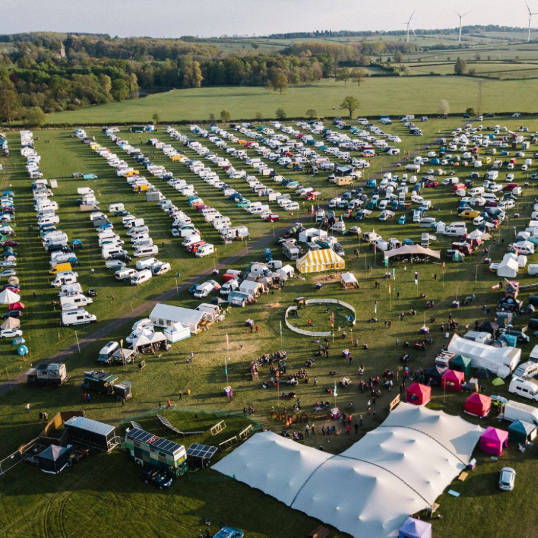 Aerial view of a large outdoor festival or event in a spacious green field. Numerous tents, RVs, and vehicles are organized in rows. Several large tents, including a striped one, are set up. People gather in various areas, and wind turbines are visible in the distance against a backdrop of trees.