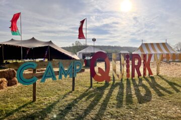 Sunlit outdoor festival scene with large colorful letters spelling "CAMP QUIRK" on the grass. Tents with bunting and flags are in the background, including a yellow-striped tent and a larger black tent. The sky is blue, slightly cloudy, and the sun is low, casting long shadows.